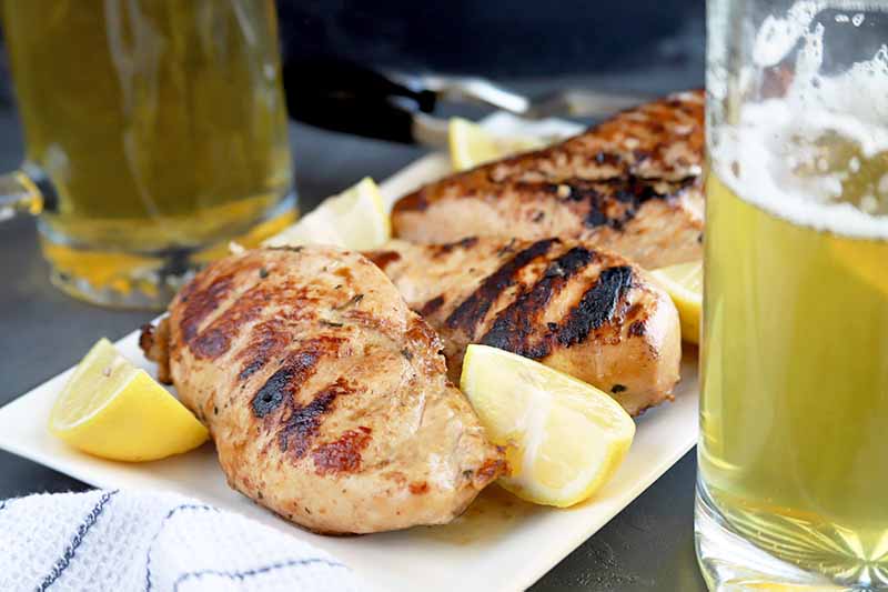Horizontal image of four grilled chicken breasts on a white serving platter with wedges of lemon, with two mugs of beer to the right and left, and a white cloth with blue stripes in the foreground, on a gray slate surface.
