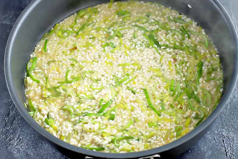 Horizontal of a large nonstick frying pan of rice, broth, sliced leeks, and other ingredients to make a vegetable risotto, on a gray slate background.