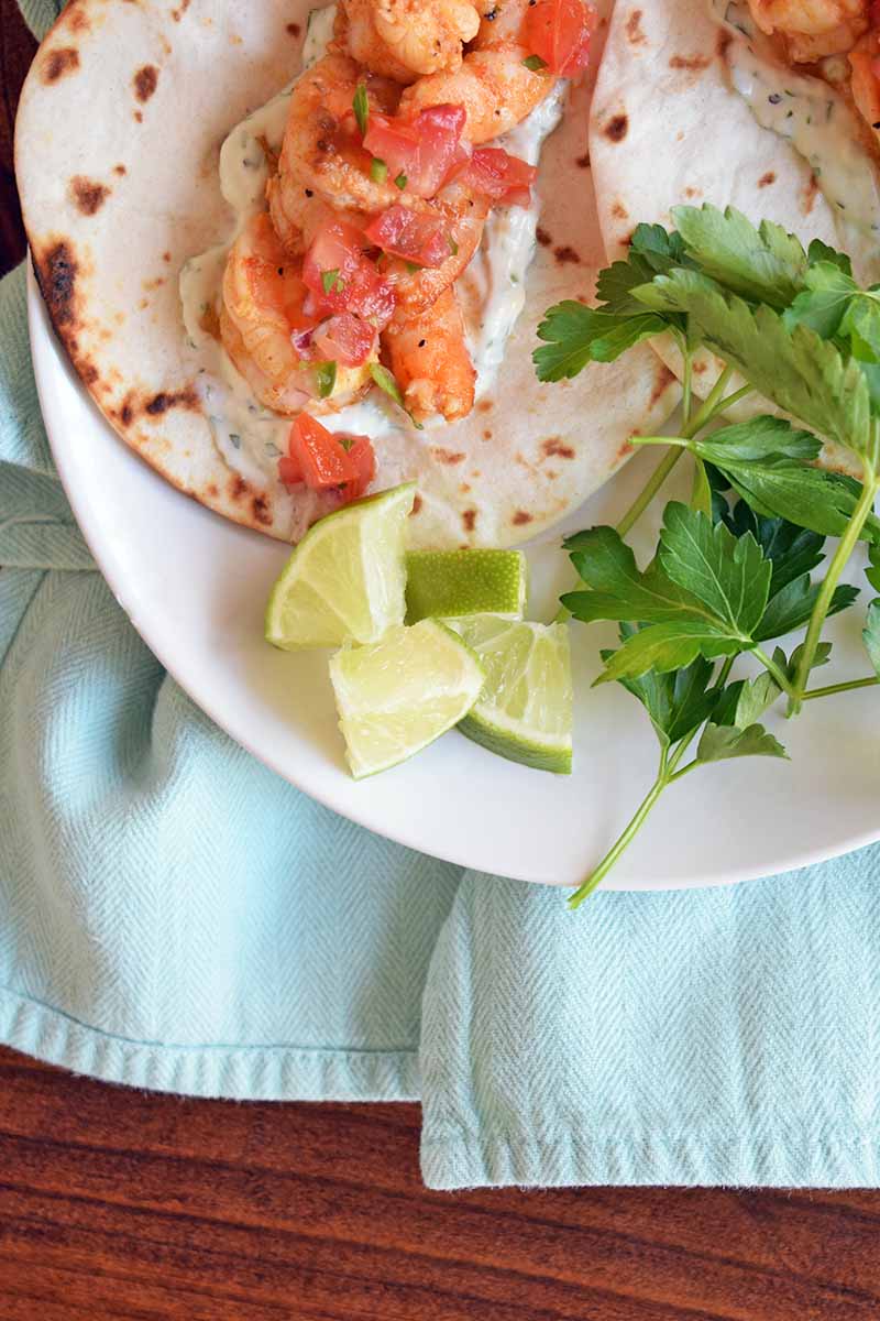 Overhead horizontal image of a white plate of shrimp tacos with a sprig of fresh cilantro and lime wedges, on a brown wood table with a folded light blue cloth napkin.