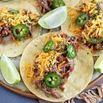 Overhead horizontal image of three beef barbacoa tacos on a beige plate, with shredded cheese, chopped cilantro, sliced jalapeno, and wedges of lime, on a striped cloth with fringe on top of a gray surface.