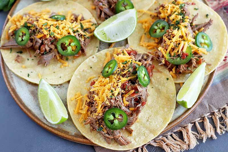 Overhead horizontal image of three beef barbacoa tacos on a beige plate, with shredded cheese, chopped cilantro, sliced jalapeno, and wedges of lime, on a striped cloth with fringe on top of a gray surface.