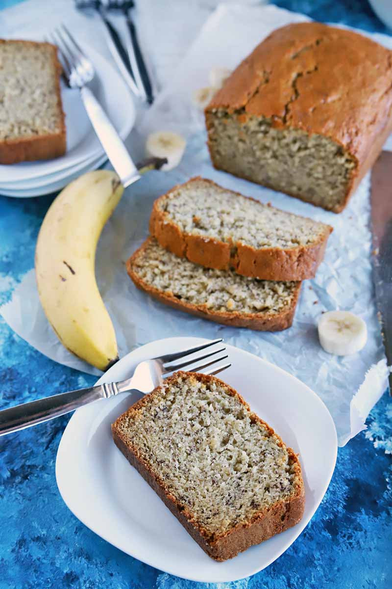 Vertical oblique overhead shot of banana bread on two plates and a white piece of parchment paper, with whole and sliced fruit and forks, on a dark and light blue surface.