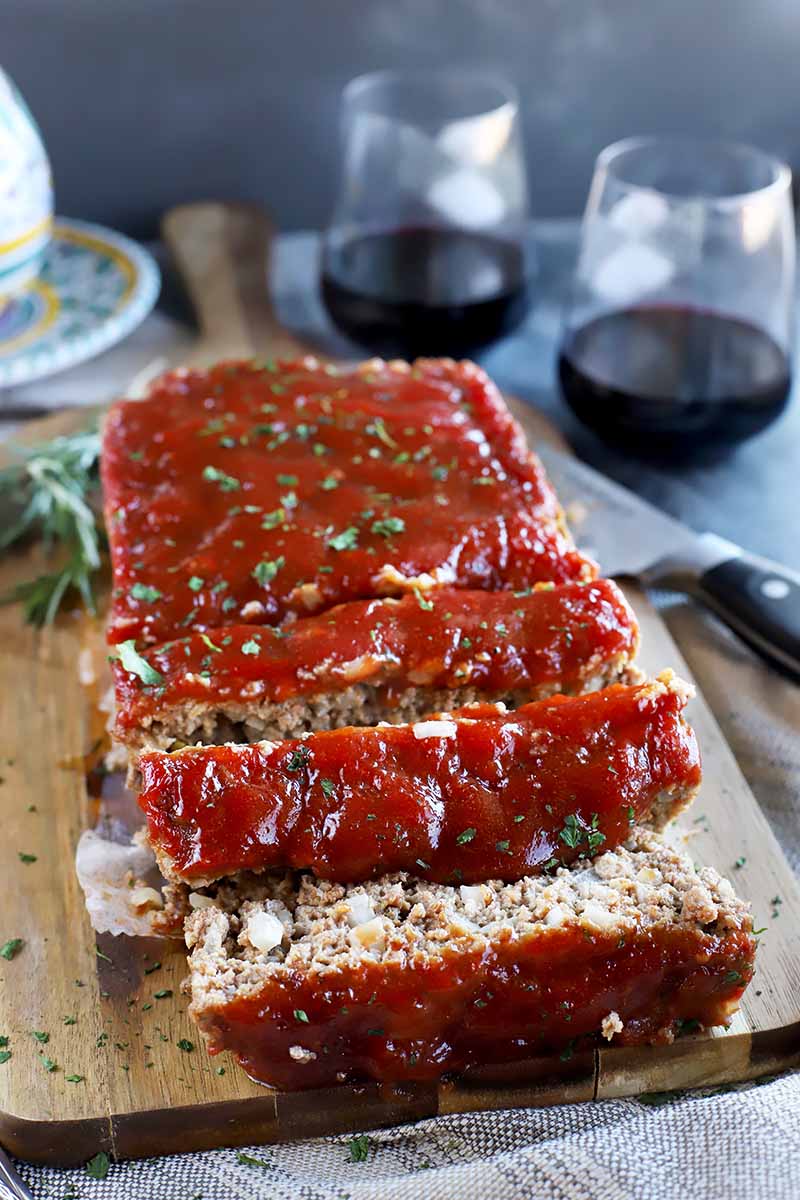 Vertical image of meatloaf partially sliced and topped with a ketchup glaze on a wooden board in front of glasses of wine.