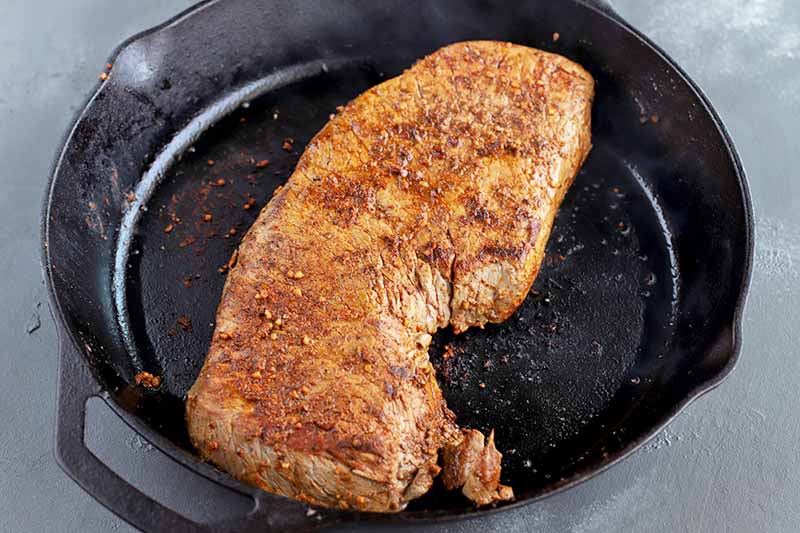 Seared seasoned beef cooking in a large cast iron pan, on a gray background.