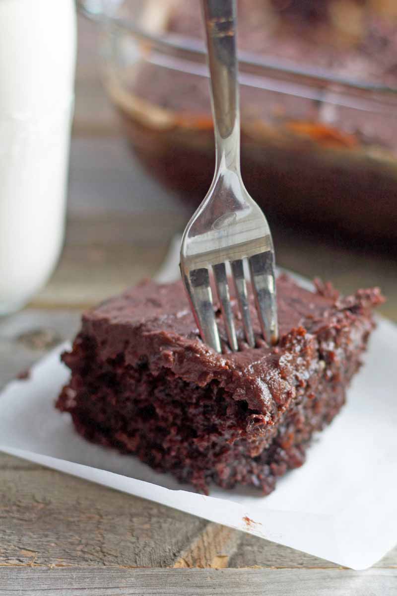 Vertical image of a fork stuck into a square of chocolate cake with chocolate frosting on a small slip of white parchment paper, on a stone surface with a bottle of milk and a glass baking dish of more of the dessert in soft focus in the background.