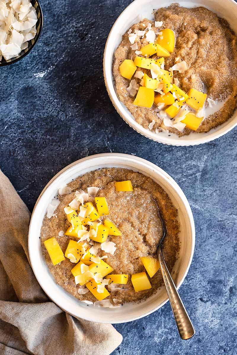 Vertical image of two breakfast bowls topped with fresh mango pieces and large coconut shreds, with one bowl having a metal spoon inserted into it.