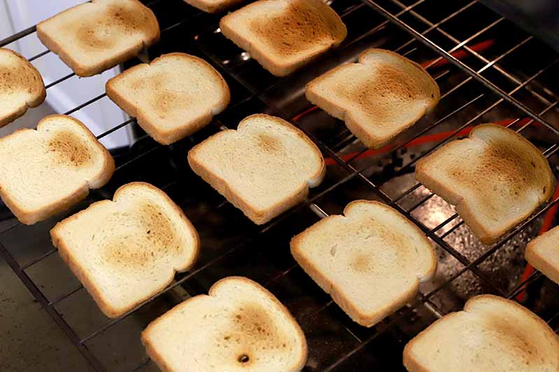 Vertical image of white bread slices with different portions that are toasted golden brown, in diagonal rows on a baking rack in an oven.