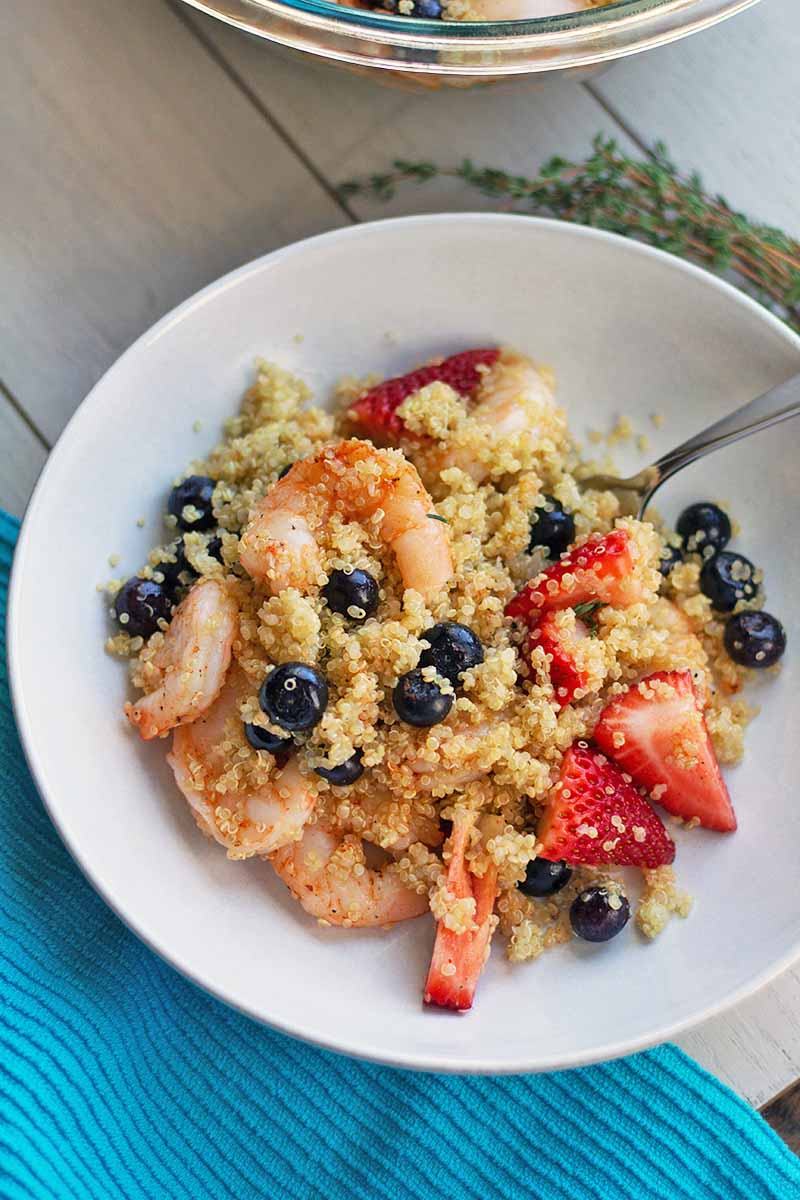 Overhead vertical image of a white bowl of quinoa with fresh berries, shrimp, honey, and seasonings, on an off-white wood surface with sprigs of thyme and a blue cloth.