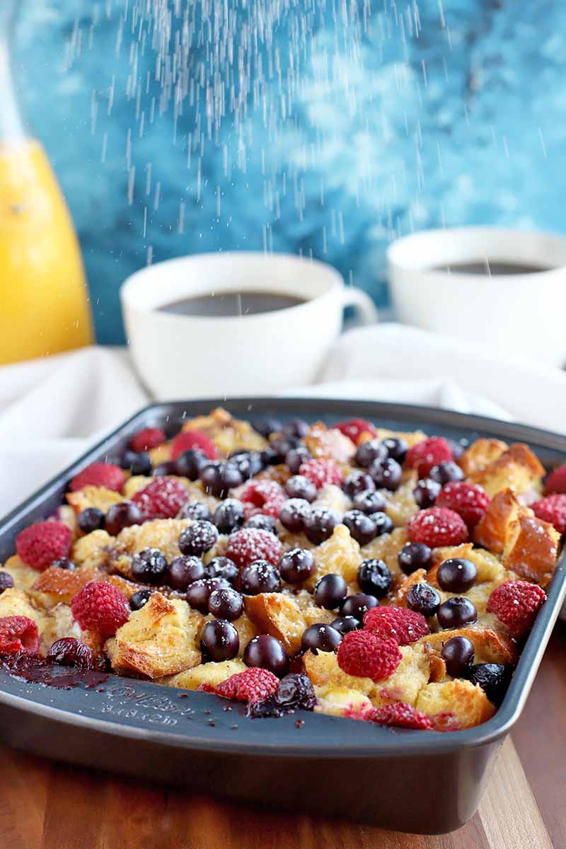 Vertical nearly head-on image of a metal baking pan filled with a fresh fruit breakfast casserole, with a glass bottle of juice and two white teacups of coffee on a white cloth in the background, against a mottled blue and white backdrop.