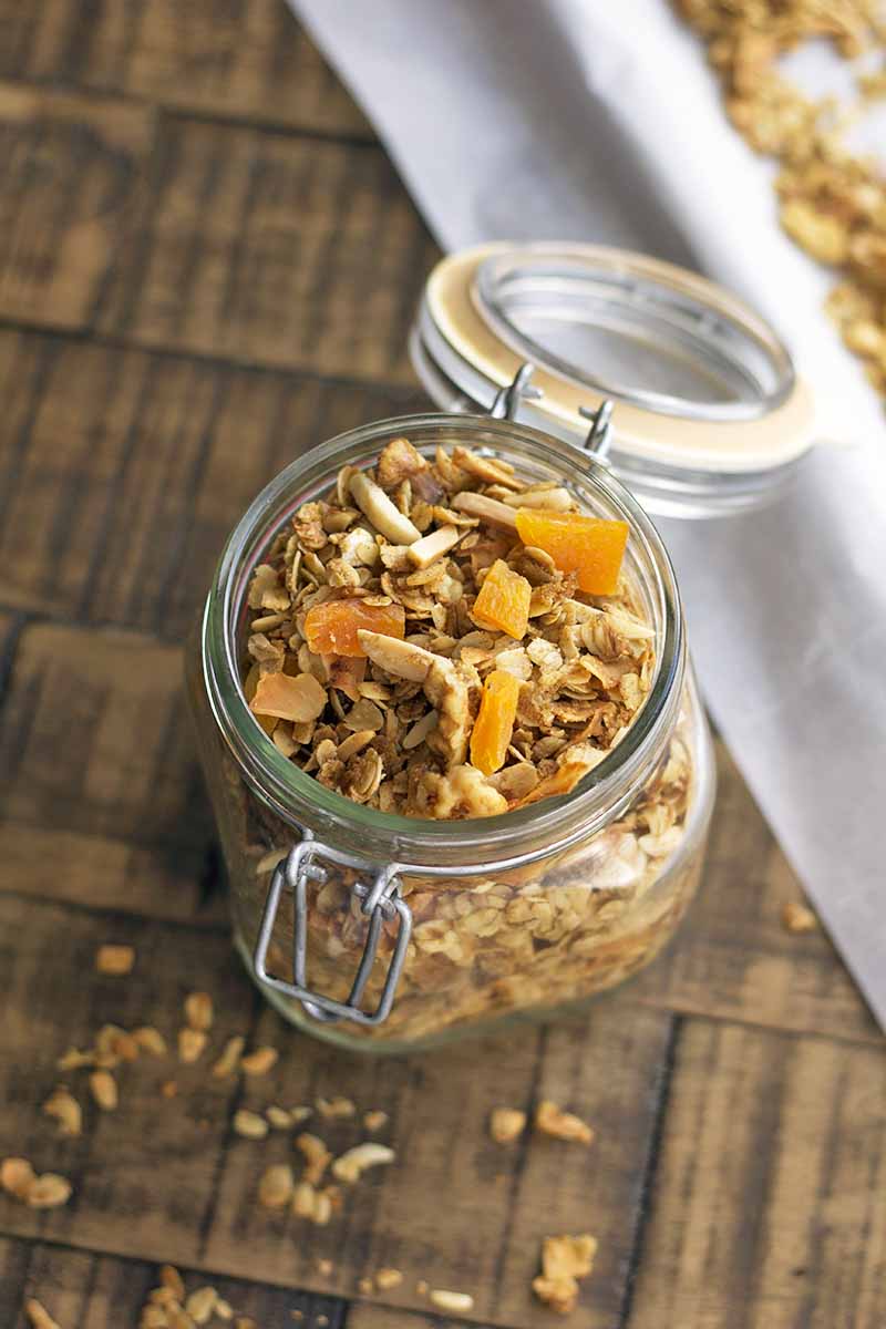 Overhead oblique image of a glass swingtop jar of homemade granola with chunks of dried apricots and chopped nuts, on a brown surface with scattered crumbs of the cereal, and a baking sheet topped with parchment and more of the breakfast food to the right.
