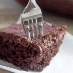 Horizontal image of a fork stuck into a square of chocolate cake on a white piece of parchment paper, on a gray surface with a bottle of milk and a glass baking dish in the background.