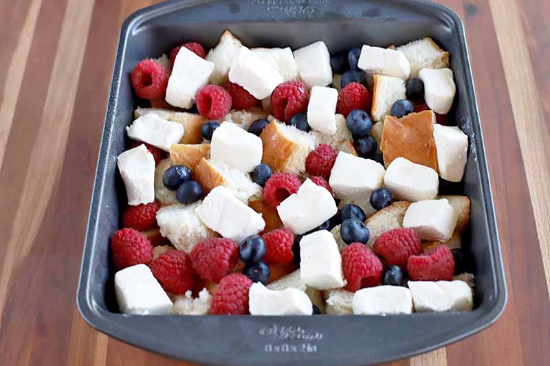 Cubes of cream cheese and fresh raspberries and blueberries are on top of cubes of crusty bread in a metal baking pan, on a wood surface with vertical stripes.