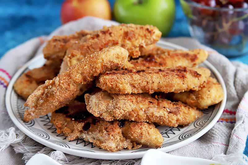 Horizontal image of a plate of stacked chicken fingers on a towel with apples and grapes.