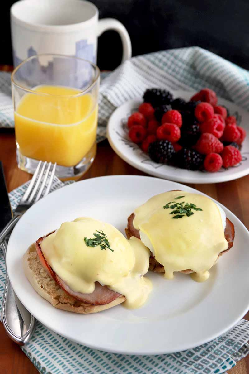 Vertical overhead slightly oblique image of eggs Benedict on a white plate with a folded blue and white checkered napkin and silverware, a glass or orange juice, a mug, and a bowl of fresh berries, on a brown surface with a black background.