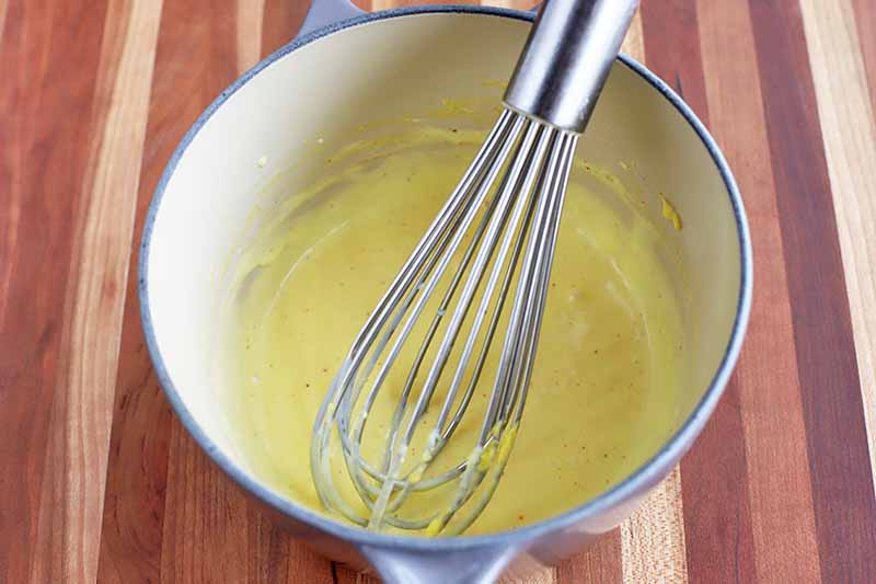 Horizontal overhead image of pale yellow hollandaise sauce being stirred with a whisk in a small saucepan, on a brown striped wood surface.