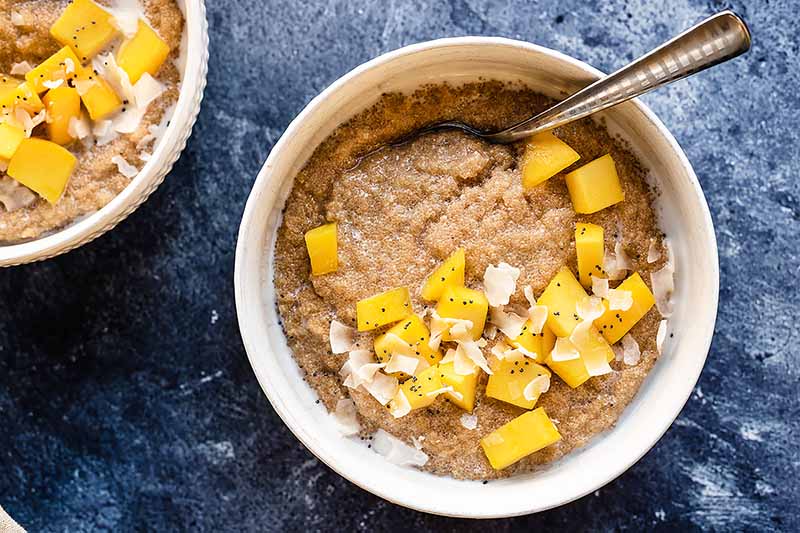 Horizontal image of breakfast bowls garnished with fresh mango chunks, coconut shreds, and chia seeds on a dark blue surface.