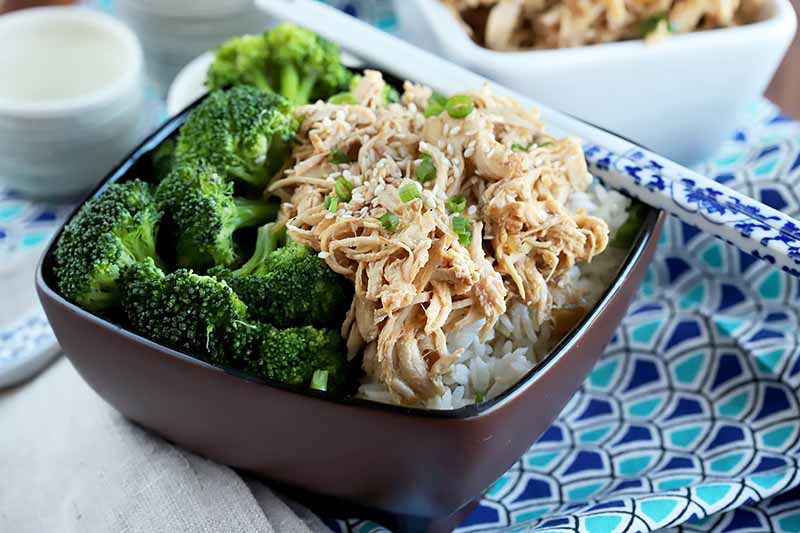 Horizontal image of a brown dish with broccoli florets and shredded chicken with white chopsticks.