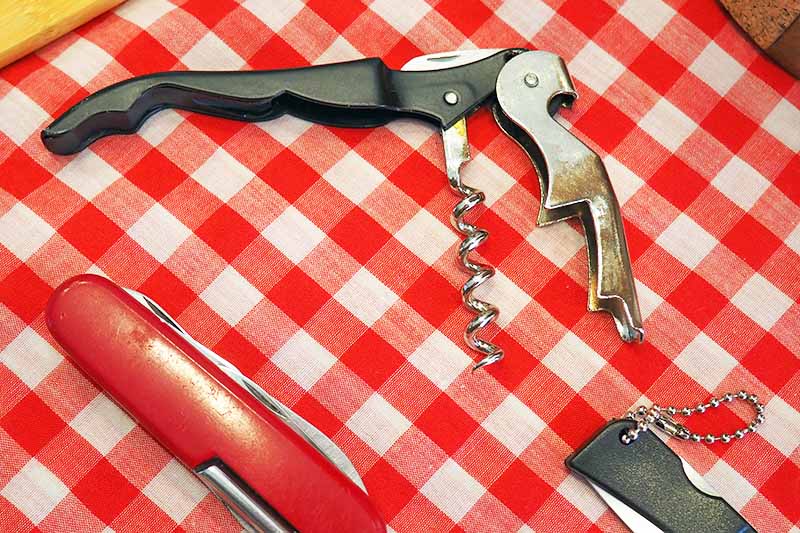 A corkscrew and two folding knives sit on top of a checkered picnic blanket.
