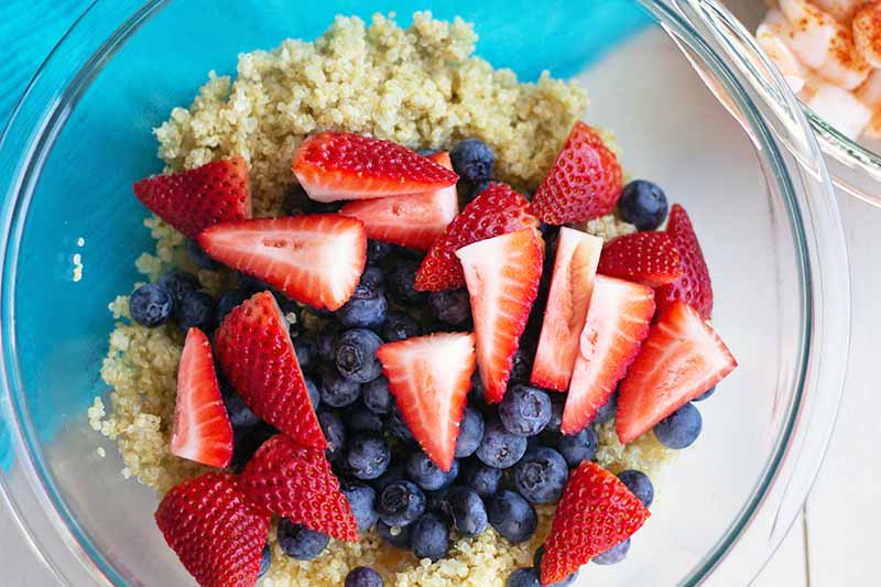 Overhead horizontal image of sliced and whole fresh berries on top of cooked quinoa in a glass mixing bowl, with a smaller bowl of cooked shrimp just barely visible at the top left corner of the frame, on a white surface partially covered at the top left with a bright blue cloth with a ribbed texture.