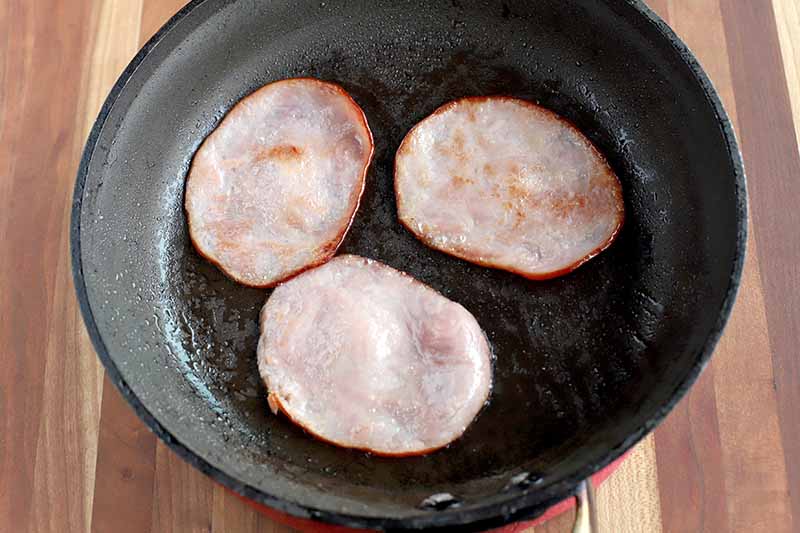 Sliced Canadian ham is frying in a nonstick saucepan, on a wood background.
