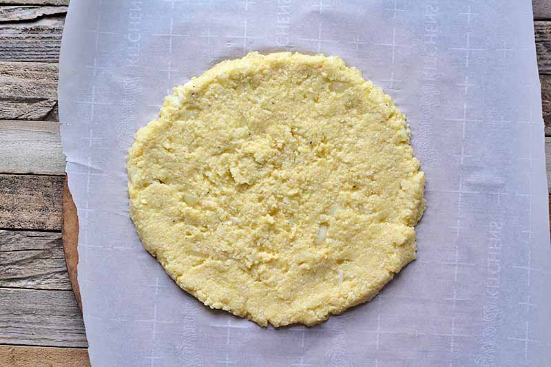 Horizontal image of an unbaked cauliflower pizza crust on a pizza stone lined with parchment paper.