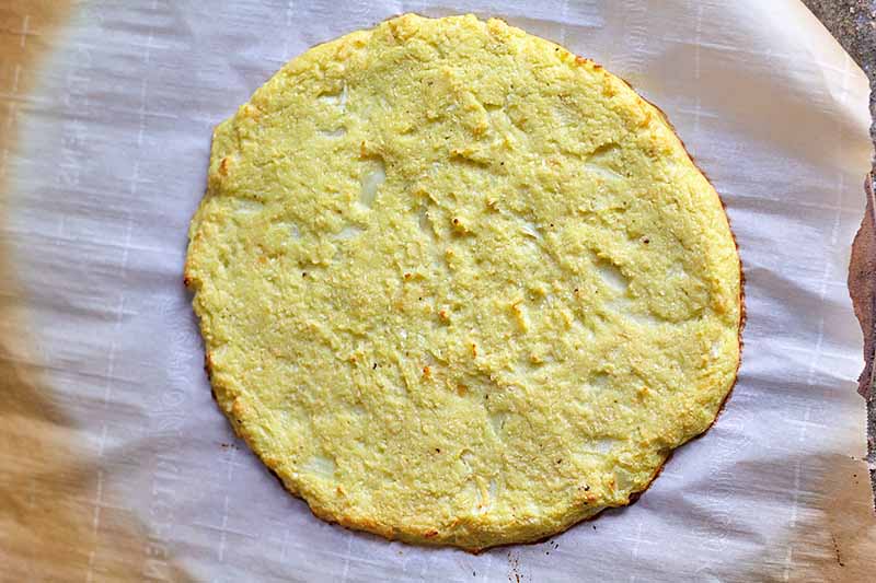 Horizontal image of a baked plain cauliflower pizza crust on a stone lined with parchment paper.