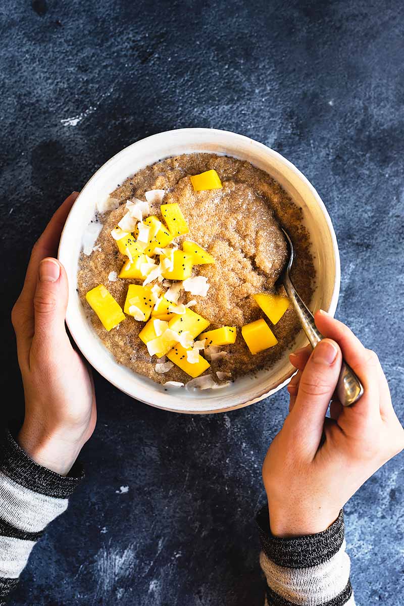 Vertical image of hands holding a spoon and a bowl of amaranth porridge with mango chunks and coconut shreds as garnishes.