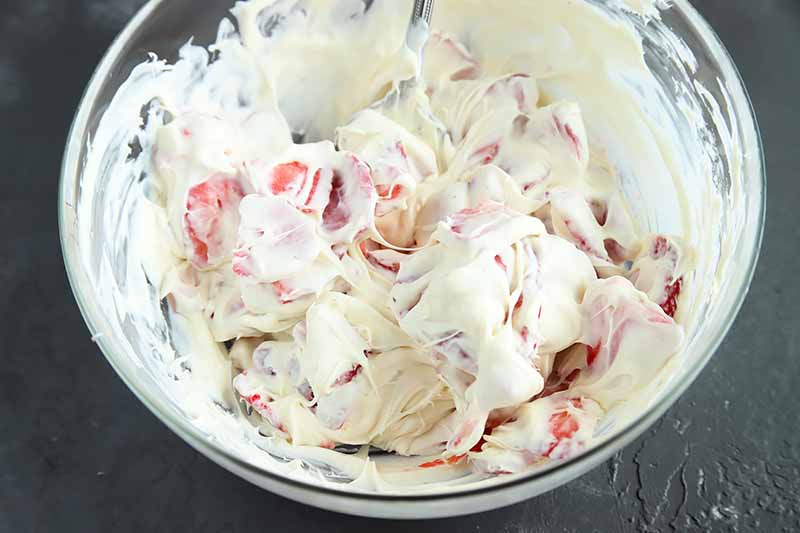 Horizontal image of strawberries mixed into a creamy white filling in a glass bowl.
