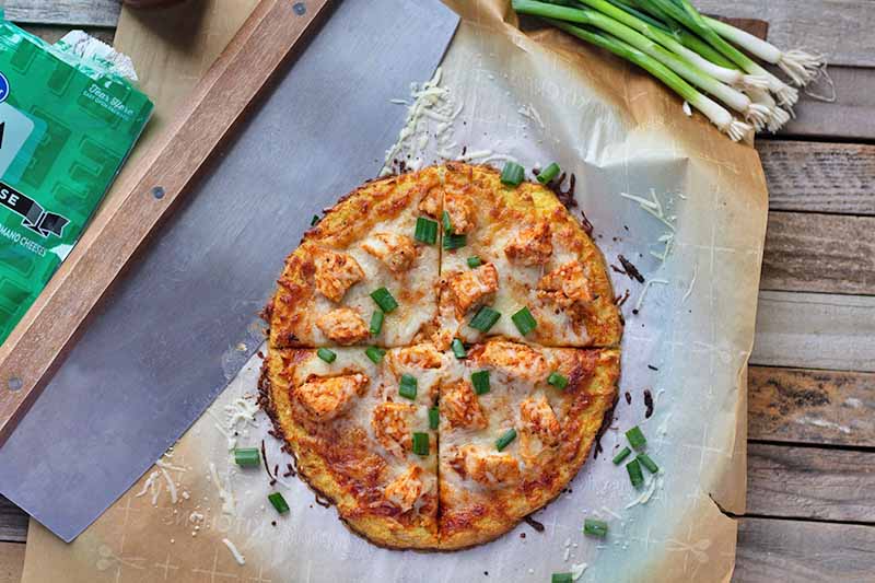Horizontal image of a whole pizza with buffalo chicken, cheese, and green onions divided into four pieces next to a mezzaluna.