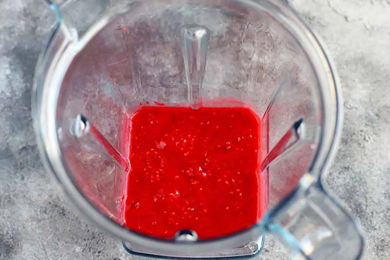 Overhead image of a plastic countertop blender with red berry puree at the bottom, on a gray and white surface.