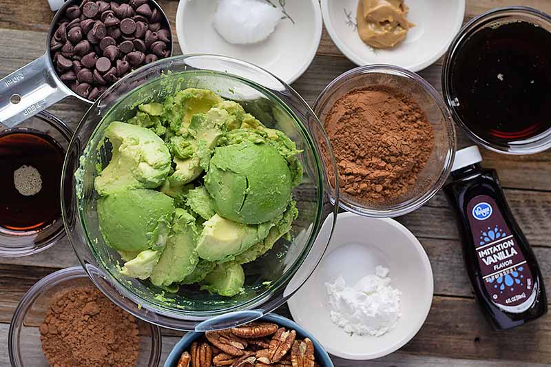 Horizontal image of a big bowl of avocado next to other bowls of ingredients on a wooden table.