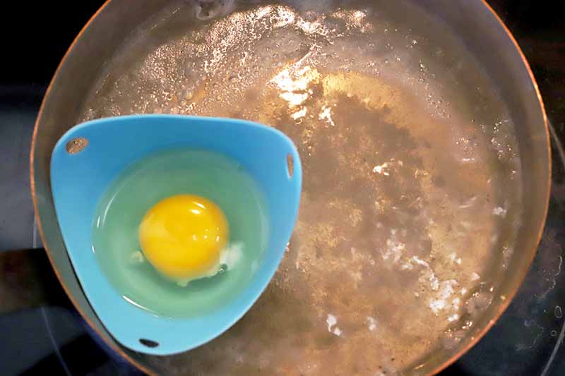 A cracked egg in a small blue silicone cup, floating in boiling water in a saucepot.
