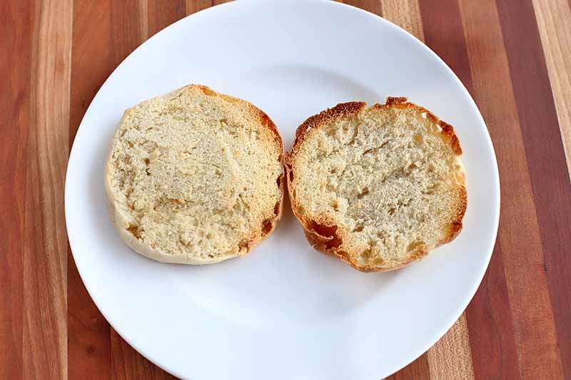 Two halves of a toasted English muffin on a white plate, on a brown wood background with vertical stripes.
