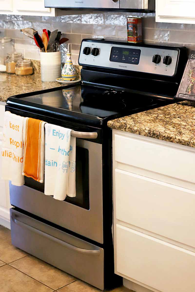 A kitchen with white cabinets and brown granite countertops, a flat-top stove, towels hanging from the oven door handle, a utensil canister with spoons and other items on the countertop, with a microwave above, and beige tile floor.
