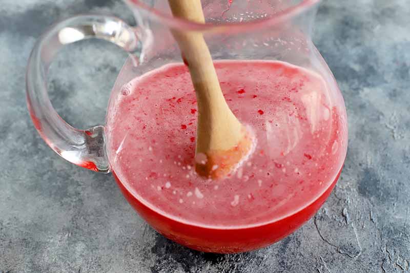 A wooden spoon stirs a foamy red fruit mixture in a glass pitcher, on a gray surface.