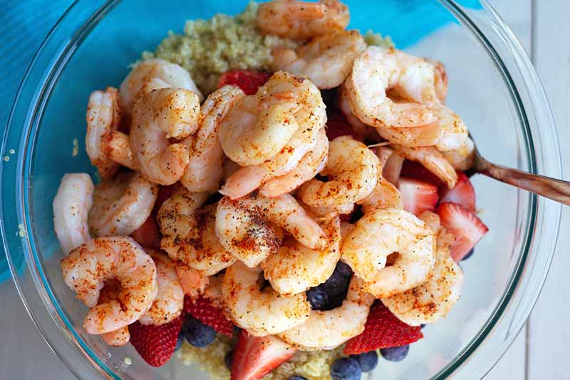 Overhead horizontal image of a large mixing bowl of cooked shrimp seasoned with paprika and other spices, fresh berries, and cooked quinoa, on a white surface covered in the top left quadrant of the frame with a blue place mat.