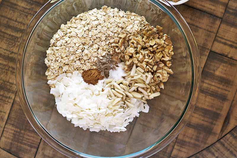 Overhead horizontal image of a large glass mixing bowl with piles of unmixed oats, nuts, coconut, and spices at the bottom, on a wood surface.