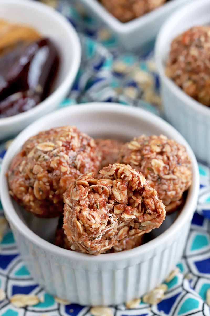 Vertical close-up image of oat balls in a white ramekin, one with a bite taken out of it.