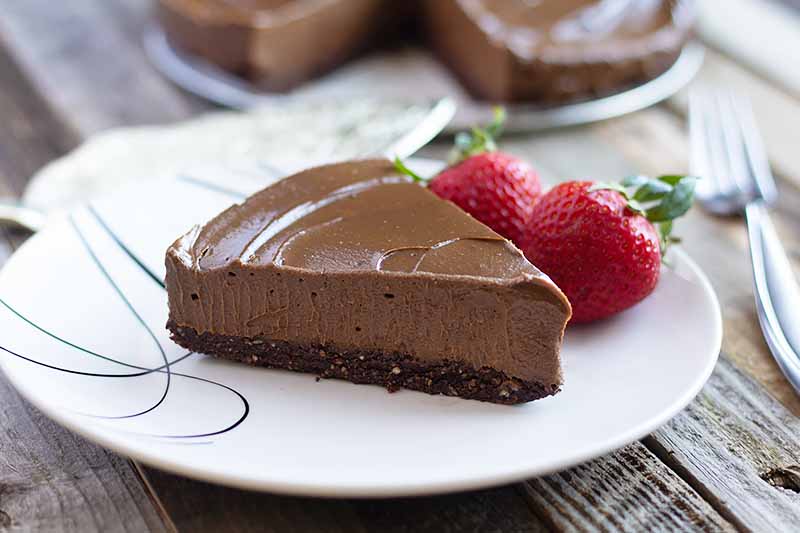 Horizontal image of a slice of a creamy chocolate torte on a white plate next to two whole strawberries.