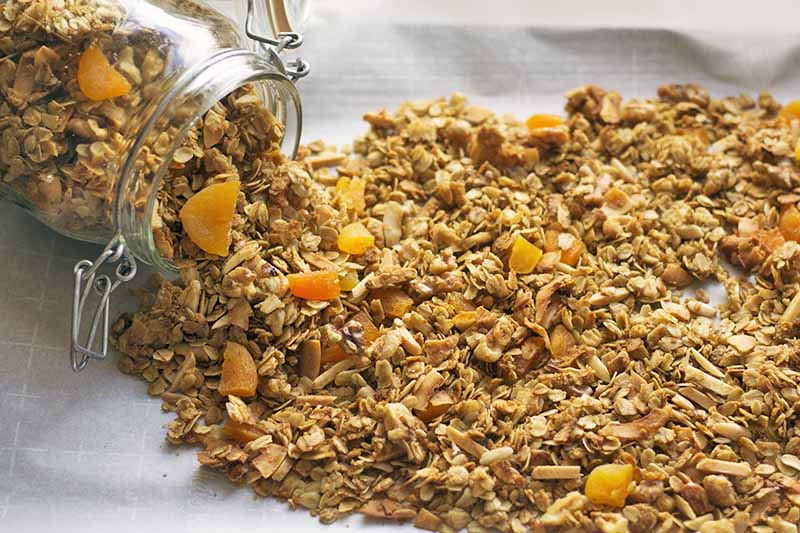 A glass swingtop jar of homemade granola is spilling onto a white parchment paper surface.