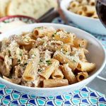 Horizontal image of a white bowl with rigatoni on a blue patterned napkin next to bread slices and a glass of red wine.