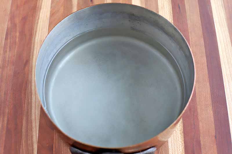 About an inch of water is in the bottom of a saucepan, with a little salt and white vinegar, on a striped wood surface.