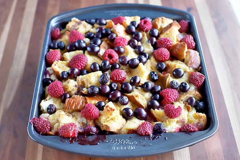 Baked fresh blueberries and raspberries with cubed bread, baking in a metal pan, on a wood surface with vertical stripes.