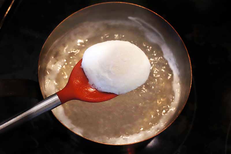 A red silicone spatula with a metal handle is being used to lift a poached egg out of a pan of simmering water, with a black background.