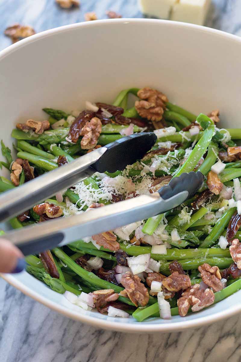 Vertical image of a hand using metal tongs to toss an asparagus salad with dried fruit and nuts, topped with grated cheese, in a large white bowl on a gray surface.