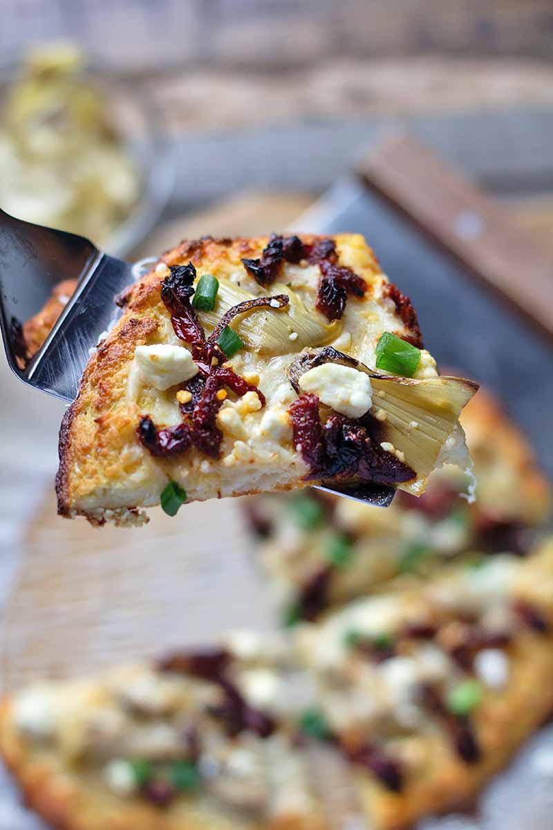 Vertical close-up image of a spatula holding a slice of cauliflower crust with green onions and sun-dried tomatoes.