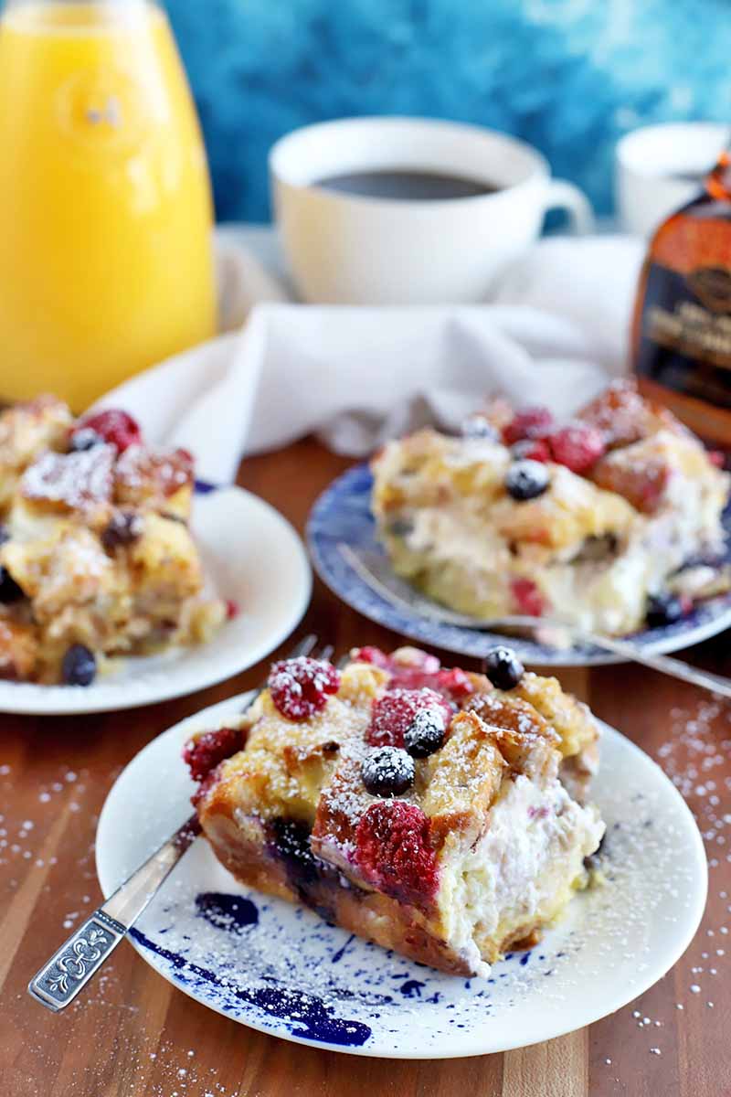 Vertical image of three small plates of berry breakfast casserole with forks, on a brown wood table, with a white cloth, cup of coffee, bottle of maple syrup, and bottle of orange juice in the background, against a mottled blue and white backdrop.