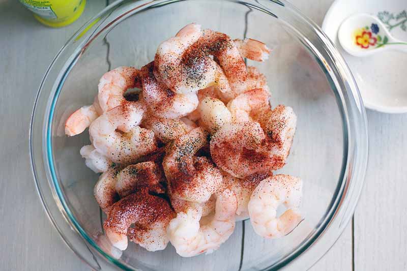 A large glass bowl of cooked shrimp with paprika and other seasonings sprinkled on top, with a ceramic dish to the left, on a white surface.
