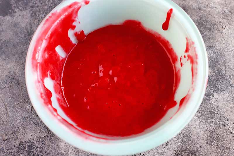 Overhead horizontal image of a white bowl of raspberry puree, on a gray background.