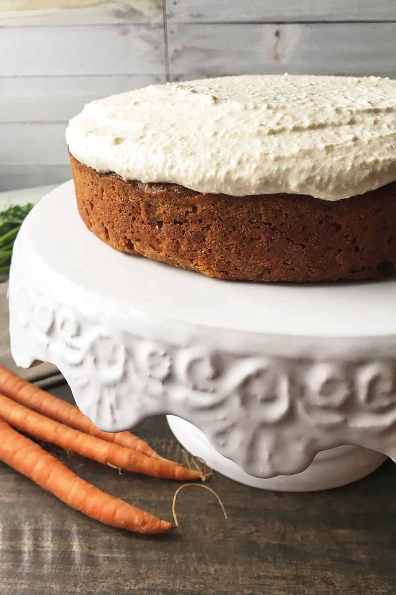 Vertical image of a one-layered orange cake topped with white icing on a white stand next to three carrots.